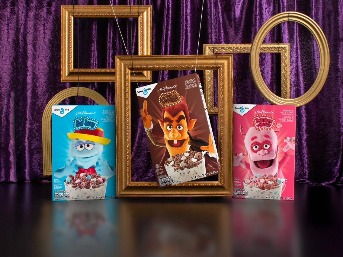 ​Three boxes of General Mills Monster Cereals — Boo-Berry, Count Chocula, and Franken Berry — are displayed on a reflective surface as if in an art gallery. The boxes, featuring Jim Henson's puppets of the characters, are set against a rich purple curtain, with empty, ornate gold picture frames hanging around them.​